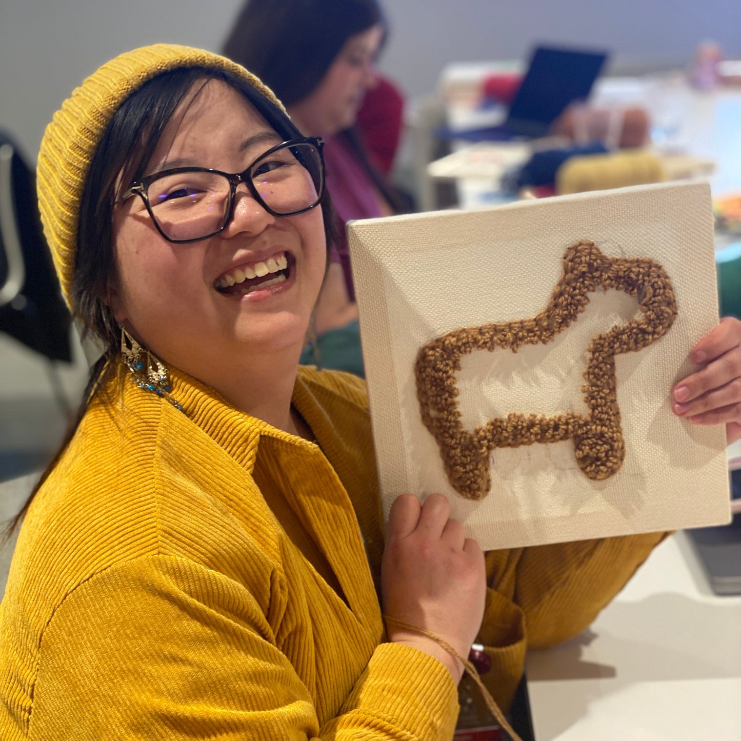 Student work from the punch needle wall hanging class. A person dressed in a yellow jacket and hat is holding up a square canvas with the outline of a capybara punched into the middle.