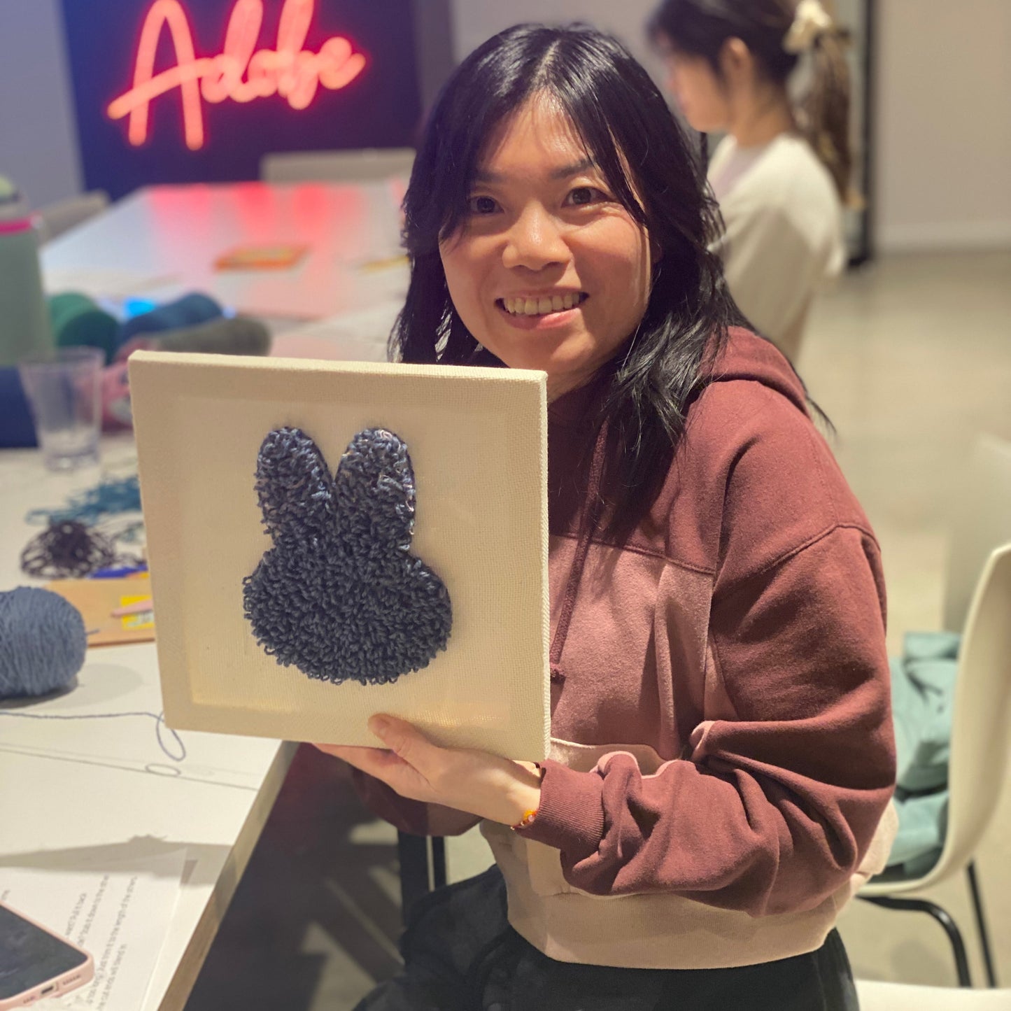 A person wearing a maroon hoodie is holding up their punch needle canvas with a gray rabbit shape in the middle.