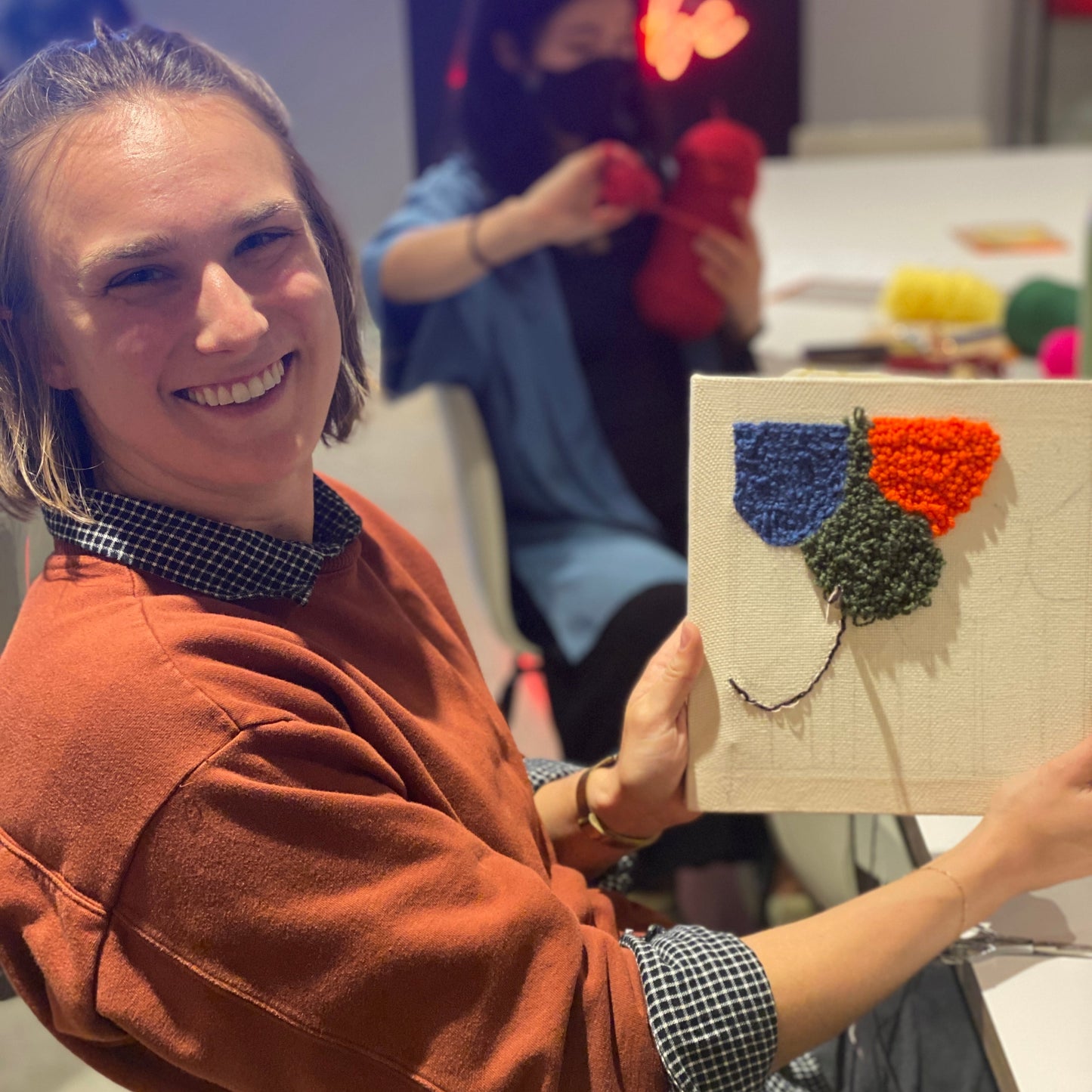 A smiling person is holding up their punch needle canvas in progress. It has three brightly colored circles at the top.