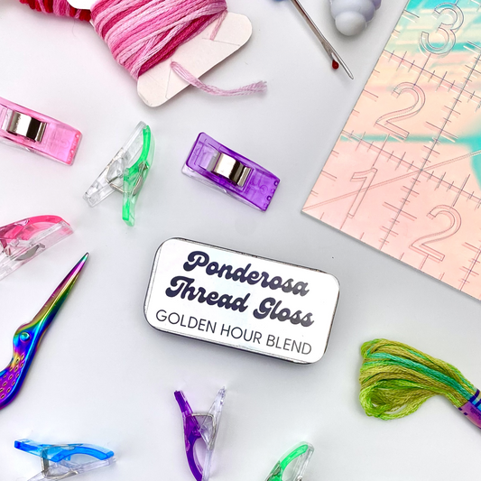 A metal tin of Ponderosa Thread Gloss - Golden Hour Blend - against a background of scattered, colorful sewing supplies including: embroidery floss, sewing clips, iridescent scissors, seam ripper and a cutting mat.