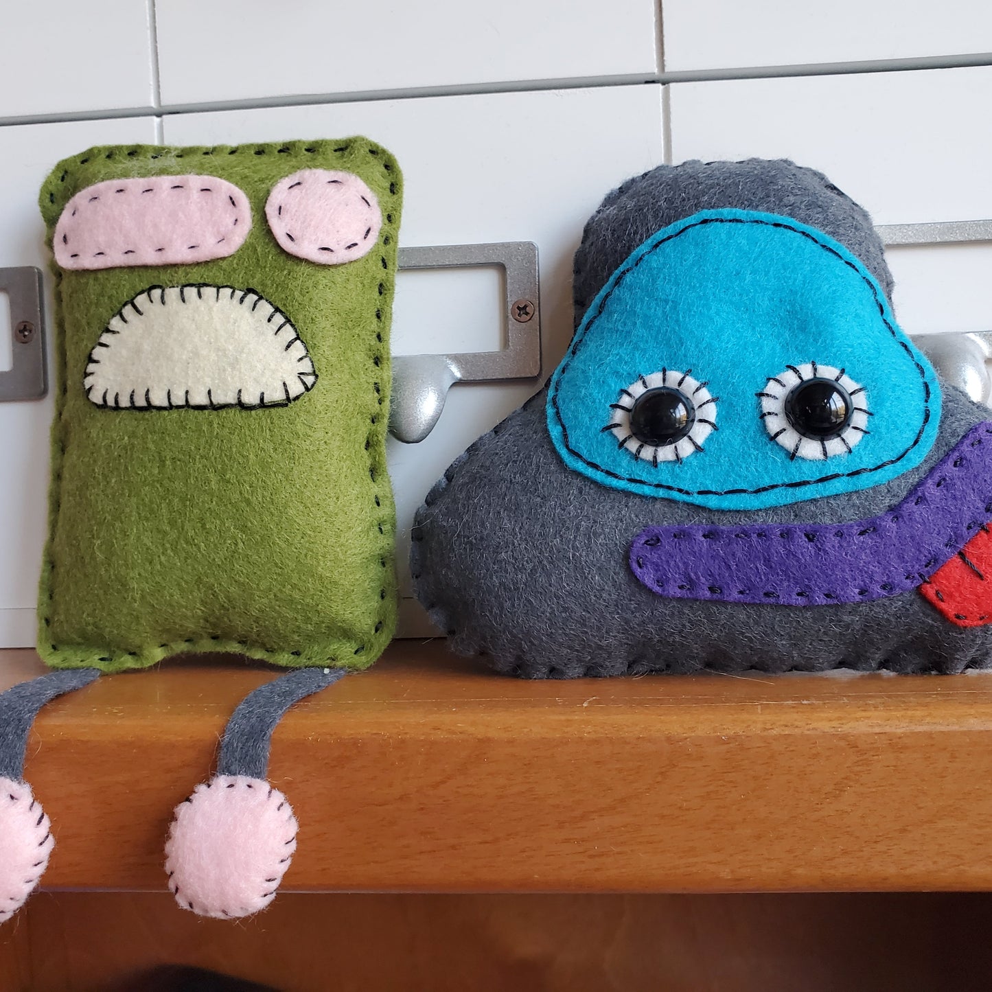 Two examples of felt monsters sitting next to each other on a shelf. One is a green rectangle with pink eyes and a downturned mouth. This one has dangly legs that dangle off the shelf. His friend is a triangle shape wearing an eye mask. It has a purple mouth with a red tongue hanging out.