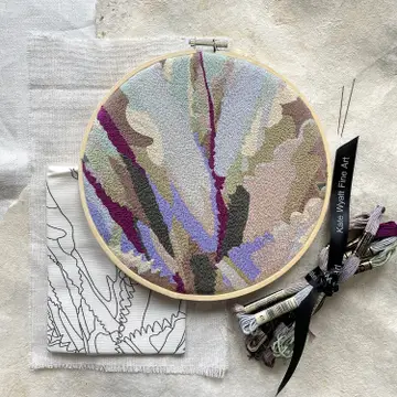 An example of the finished embroidery kit agave with blue, purple and natural colors. A cloth with the printed pattern is folded below the example.