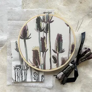 An example of the finished embroidery kit - thistle with blue, purple and natural colors. A cloth with the printed pattern is folded below the example. A batch of embroidery floss is tied with a ribbon and sitting on the table next to it.