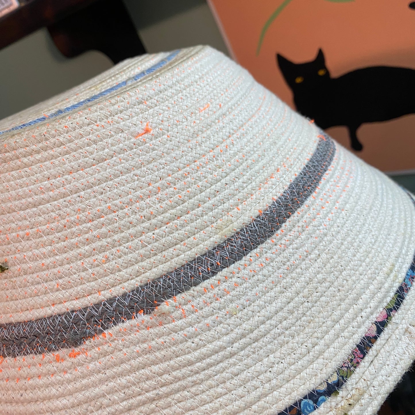 An example of a machine sewn rope bowl. The bowl is upsidedown in the photo. The stitching has a smattering of neon orange and white thread on a cream colored rope. Some gray and black fabric scraps are used for contrast.