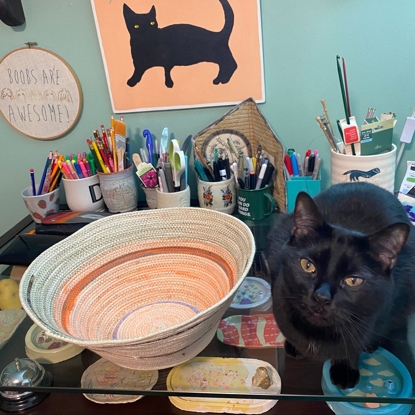 A black cat named Meat Monster has broken into the filming of this example rope bowl. She sits on the desk next to the rope bowl. A painting of her against a peach background hangs above the bowl.