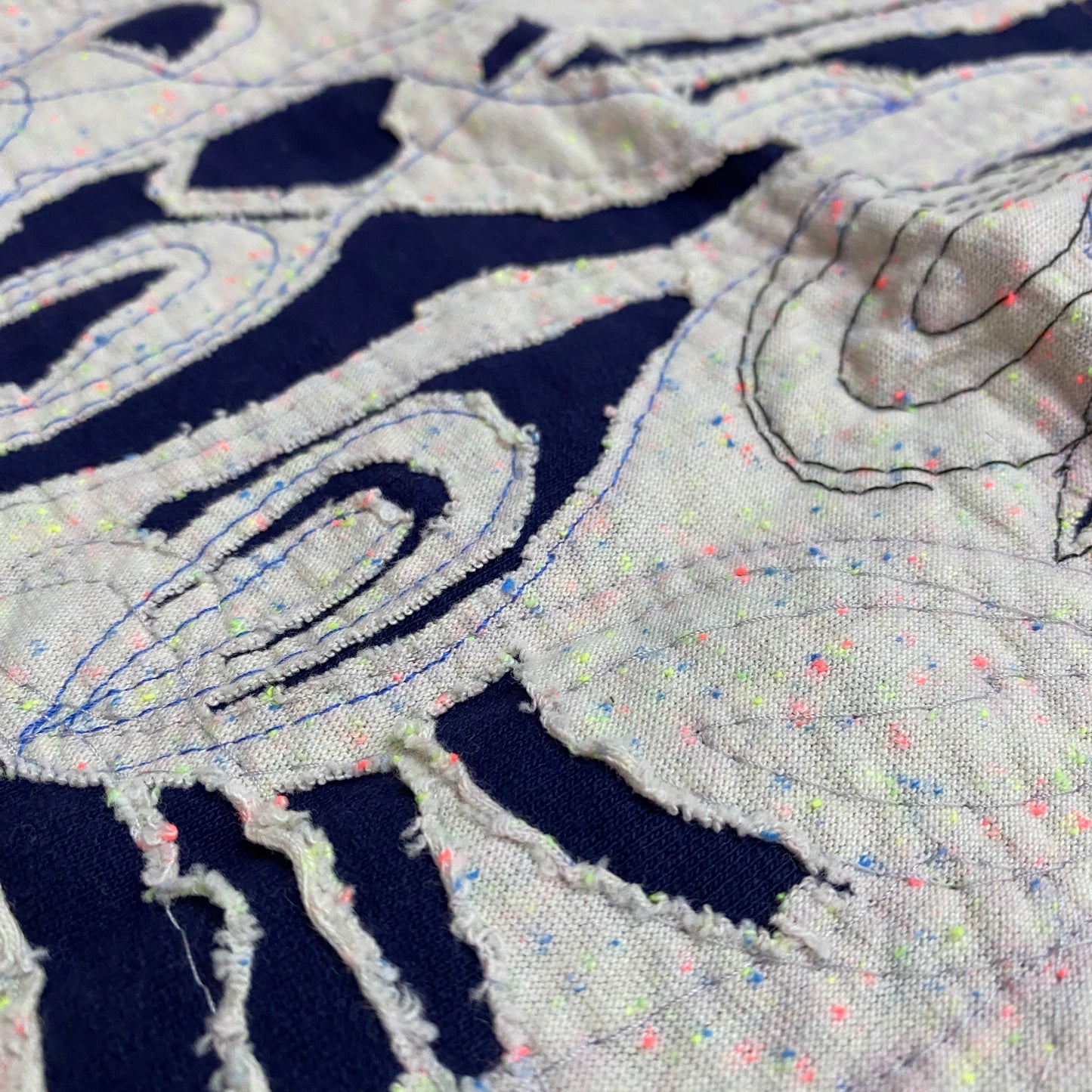 The background fabric on this negative reverse applique bandana is navy blue. The foreground fabric is cream colored with neon flecks. The top stitching is navy blue.