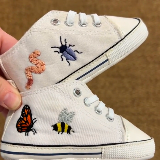 A pair of white, canvas baby shoes have been embroidered with a variety of bugs.