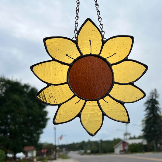 A stained glass sunflower hanging in a window by a metal chain.