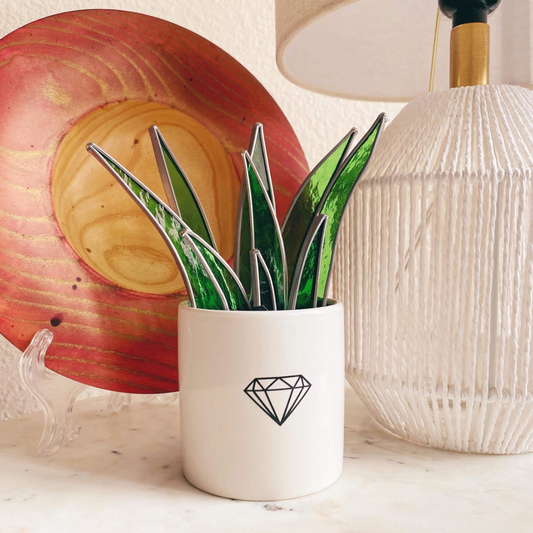 A potted plant made from stained glass sits in a white pot with a crystal on the front. A white lamps and a wooden bowl are on the table as well.