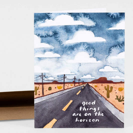 A greeting card with a painted image of a long stretch of straight highway. There are cacti and telephone poles. The sky is bright blue with big fluffy clouds. It says "Good things are on the horizon" on the right side of the road.