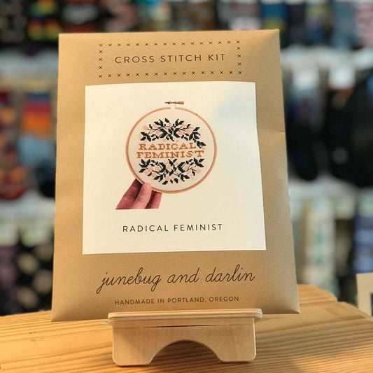 The packaging for the Radical Feminist Cross Stitch Kit by Junebug & Darlin. It is a brown kraft paper envelope with a picture of the completed kit on the front. It says Radical Feminist in the middle and is surrounded by pretty floral vines.