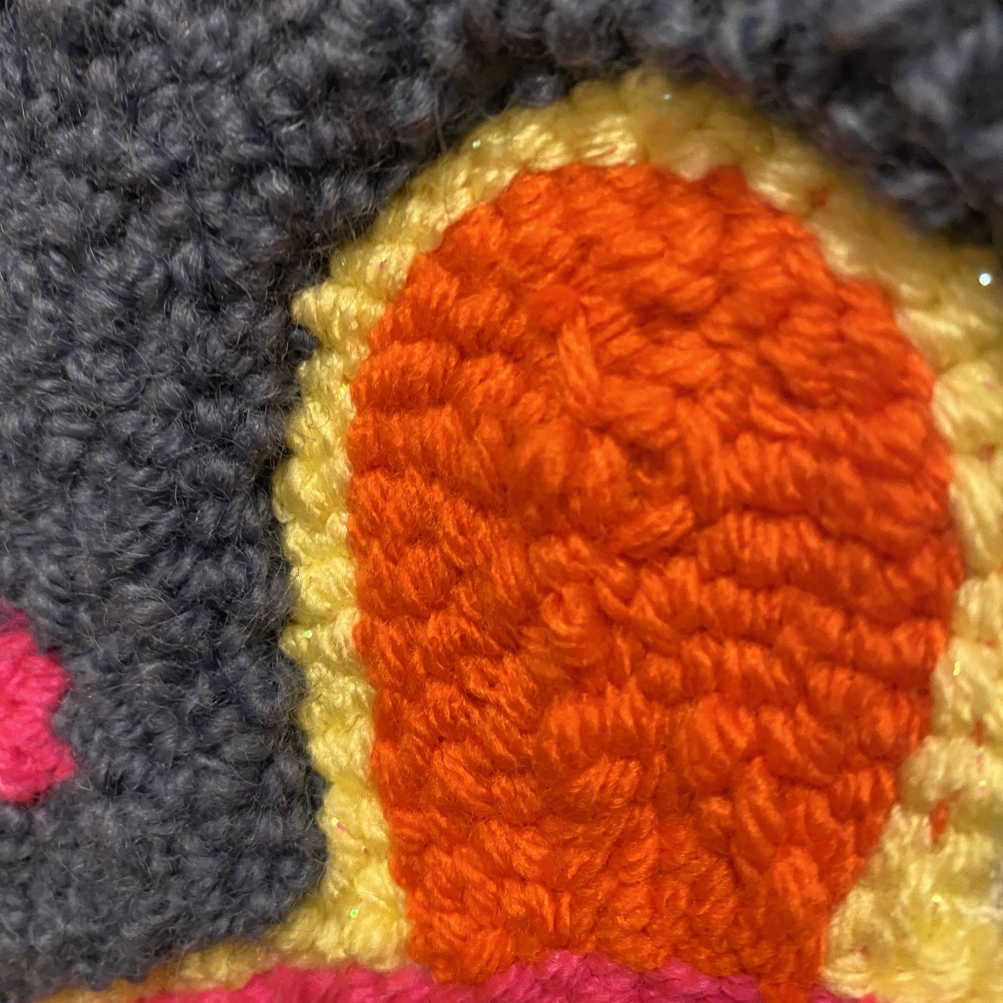 A close up view of a punch needle wall hanging. This view has two different types of stitches that are different pile heights. There is an orange balloon shape with a yellow outline and a dark purple background.