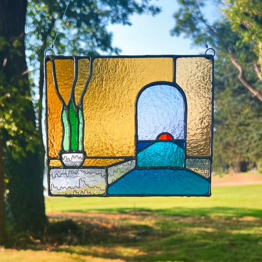 A stained glass piece with a potted plant and an arch with a sunset in the background hangs from a tree outside.