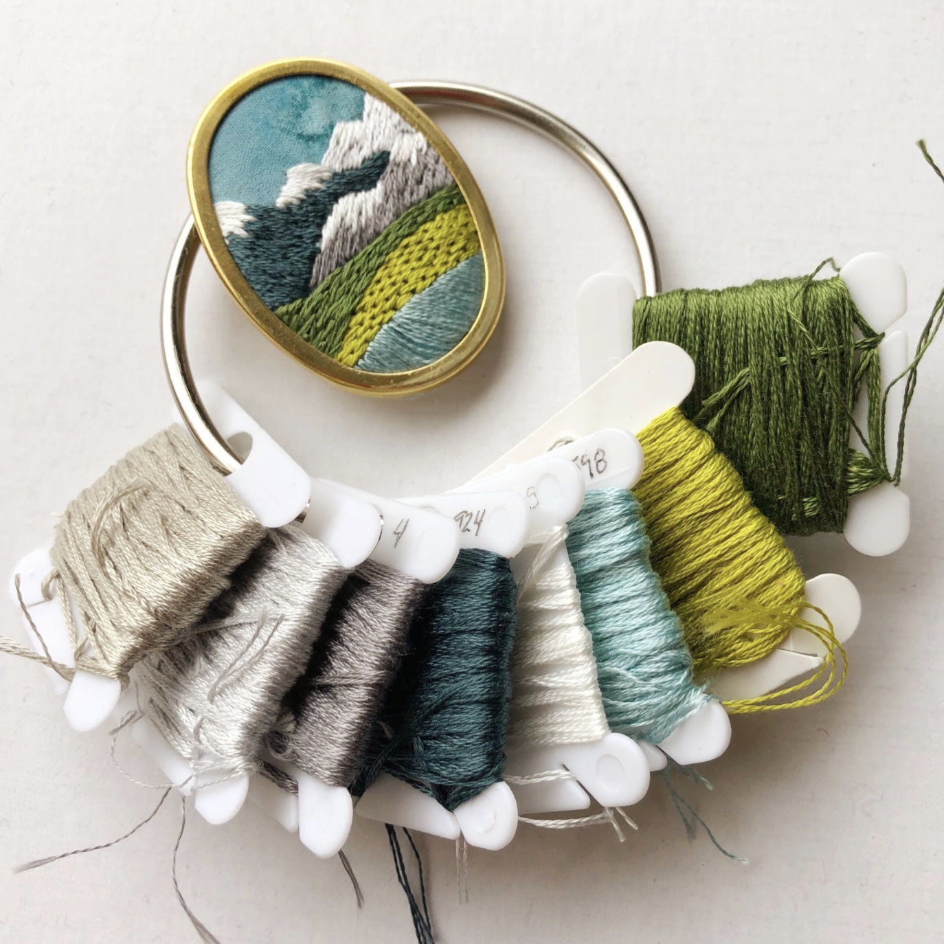 Embroidered brooch that has mountains stitched on it. It is shown next to a ring of pretty embroidery floss.