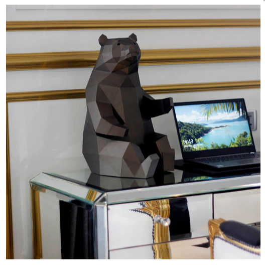 An example of a completed paper craft kit of a bear sitting down. It is on a desk next to a computer screen with a landscape screensaver.