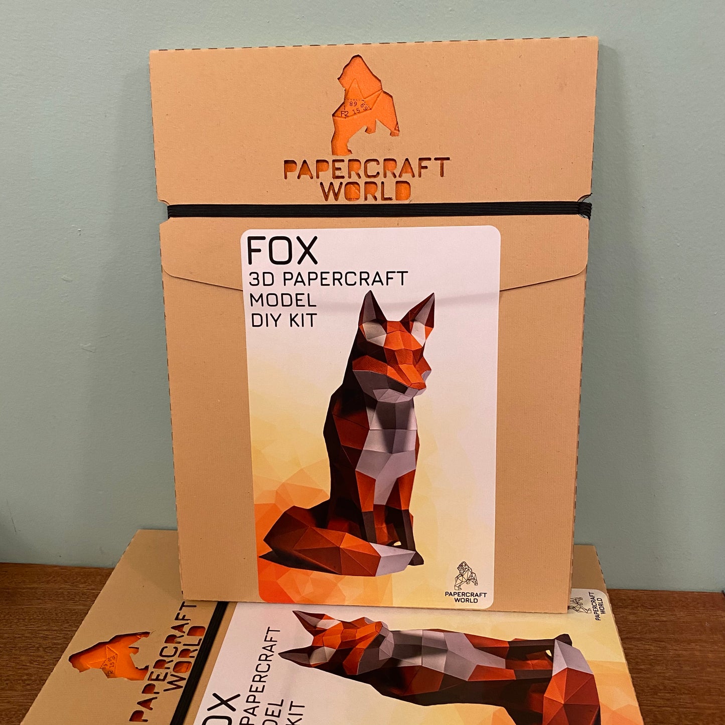 Packaging for the sitting fox paper craft kit. It is a brown paper box with the papercraft world logo laser cut into the top.
