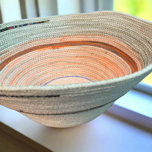 An example of a cream colored rope bowl with neon orange and olive green stitching sitting on a sunny windowsill. 