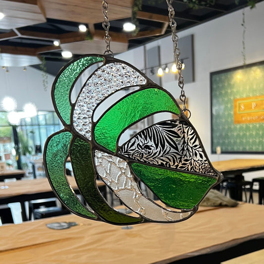 A stained glass Monstera leaf hangs from a metal chain with an empty space with tables in the background. 