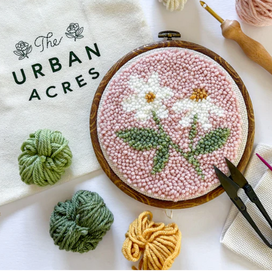A completed punch needle kit with a pink daisy is standing on a table. A cloth bag that says "The Urban Acres" is in the background. A wooden punch needle sits to the side. 
