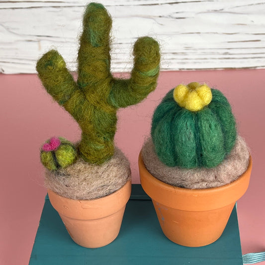 Two needle felted cacti sit on a blue wooden block. 