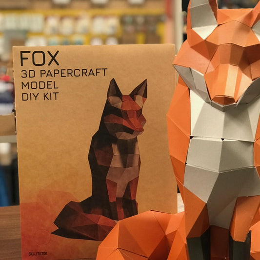 A packaged Sitting Fox Paper Craft Kit - brown paper background with an image of the completed fox on the front. An completed sitting fox sits next to the package. 
