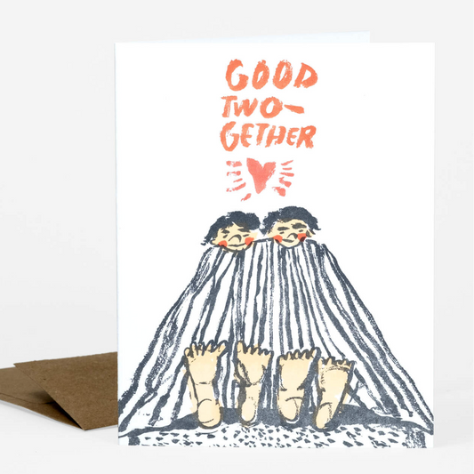 A letterpress greeting card with two dark haired people on the front. They are laying under a blanket and staring at their bare feet poking out the end It says "Good Two-gether"