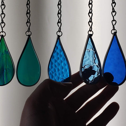 A group of stained glass raindrops hanging from metal chains. The silhouette of a hand is reaching up to touch them. 
