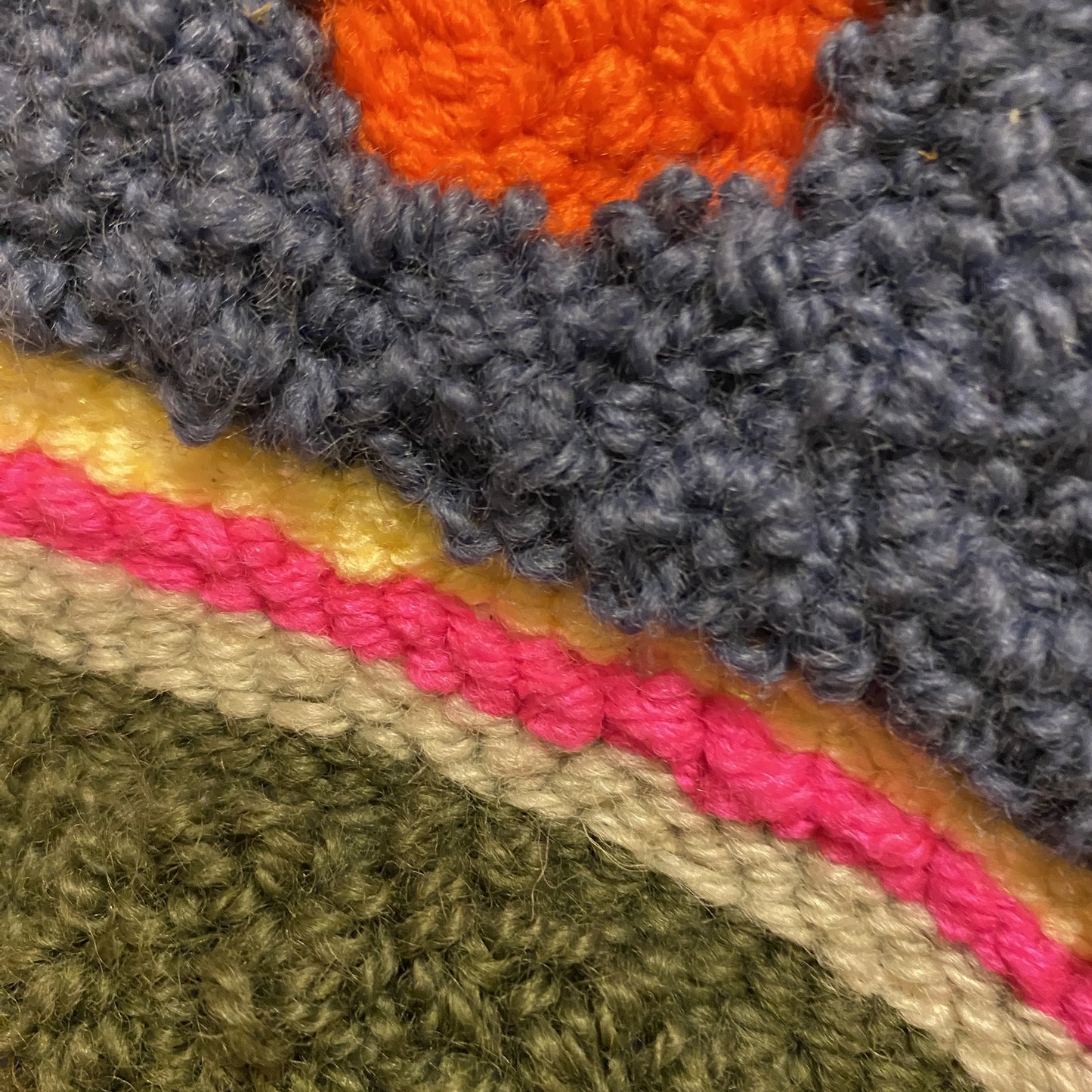 A close up view of a punch needle wall hanging. This view has two different types of stitches that are different pile heights. There are three stripes: yellow, pink and sage green with a darker green background.