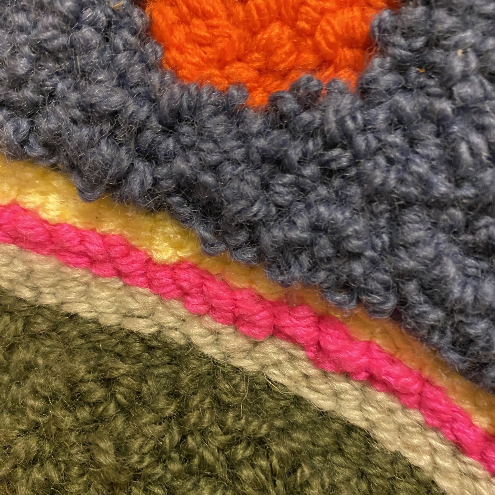 A close up view of a punch needle wall hanging. This view has two different types of stitches that are different pile heights. There are three stripes: yellow, pink and sage green with a darker green background.