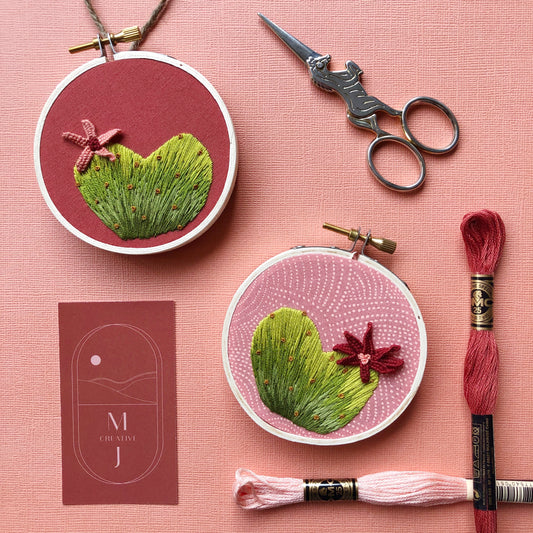 Two small embroidered pieces with pink backgrounds. A 3D heart cactus flower is embroidered at the top. A pair of metal embroidery scissors in the shape of a unicorn are in the upper right.  Two skeins of embroidery floss are in the bottom right.