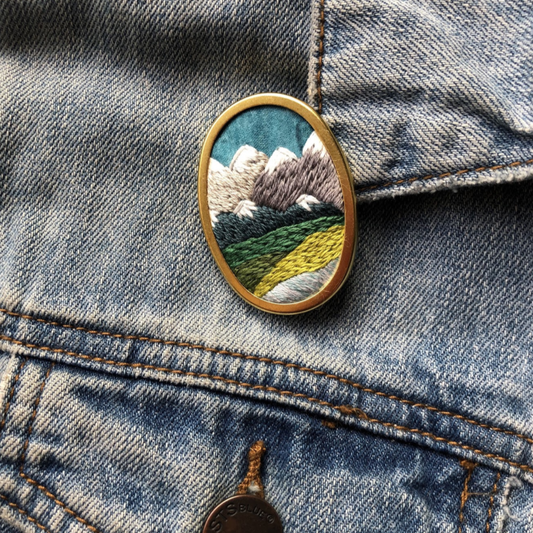 Embroidered brooch that has mountains stitched on it. It is shown pinned to a jean jacket.