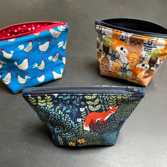 Three examples of zippered box bottom bags. On the left, chickens and roosters on a blue background and lined with red polka dot fabric & a red zipper. On the right, a Star Wars graphic lined with black fabric. In the center front, a woodland theme with pretty green foliage and a red fox. 